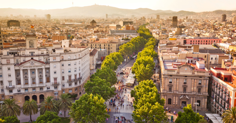 Aerial view of La Rambla, a bustling tree-lined promenade in Barcelona. The scene shows a vibrant mix of pedestrians walking along the street, surrounded by lush green trees. In the background, the cityscape is dotted with buildings, and distant hills provide a scenic backdrop. The atmosphere is lively, capturing the essence of urban life in Barcelona.