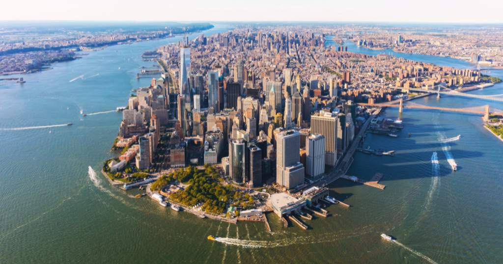 Aerial view of Manhattan, showcasing the iconic skyline surrounded by water. The image captures a bustling urban landscape filled with skyscrapers, parks, and waterfront areas. Boats can be seen navigating the waterways, while the city's grid layout is visible in the background, highlighting the vibrant energy of New York City.