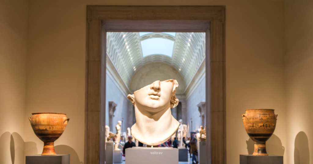 Interior view of the Metropolitan Museum of Art in New York, featuring a prominent ancient sculpture of a head on display. The sculpture is partially damaged, adding to its historical significance. In the background, a grand gallery is visible, with visitors admiring various art pieces and artifacts, illuminated by natural light streaming through the high ceilings.