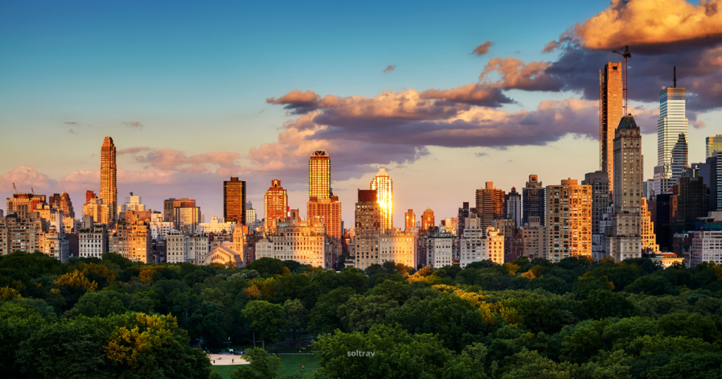 Panoramic view of the Upper East Side skyline in New York City, showcasing a vibrant sunset over Central Park. The skyline features a mix of modern and classic skyscrapers, bathed in warm golden light, with fluffy clouds scattered across the sky. Lush green trees of Central Park are visible in the foreground, creating a striking contrast against the cityscape.