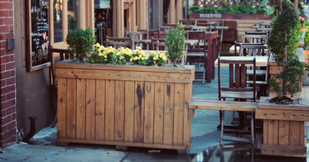 View of an outdoor restaurant in New York, featuring wooden tables and chairs arranged neatly along the sidewalk. A planter filled with vibrant yellow flowers adds a splash of color to the setting. The rustic wooden decor and the inviting atmosphere suggest a cozy dining experience, with sunlight illuminating the area and a few reflections on the pavement.