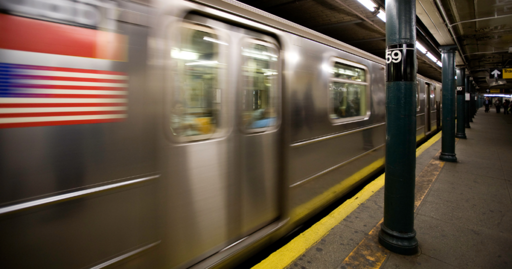 View of a moving subway train in New York City, captured in motion with a blurred effect that emphasizes speed. The train features a flag design on its side. In the foreground, a platform is visible with a numbered column indicating the station, while passengers wait nearby. The underground setting showcases the typical urban subway environment with concrete pillars and tiled walls.