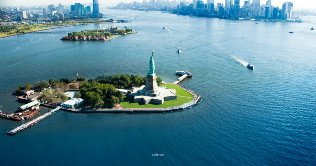 Aerial view of Liberty Island, home to the Statue of Liberty, surrounded by shimmering waters. The statue stands prominently on its pedestal, with green trees and pathways visible on the island. In the background, the skyline of New York City is visible, along with boats traversing the waterways, suggesting ferry access to this iconic landmark.