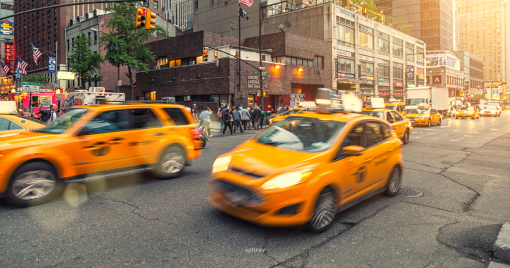 Busy city street scene featuring the iconic yellow taxis of New York City in motion. The blurred movement of the taxis suggests a vibrant, bustling atmosphere, while pedestrians cross the street in the background. The urban landscape includes buildings and traffic signals, highlighting the dynamic nature of city life. A warm glow from the setting sun adds to the inviting ambiance.