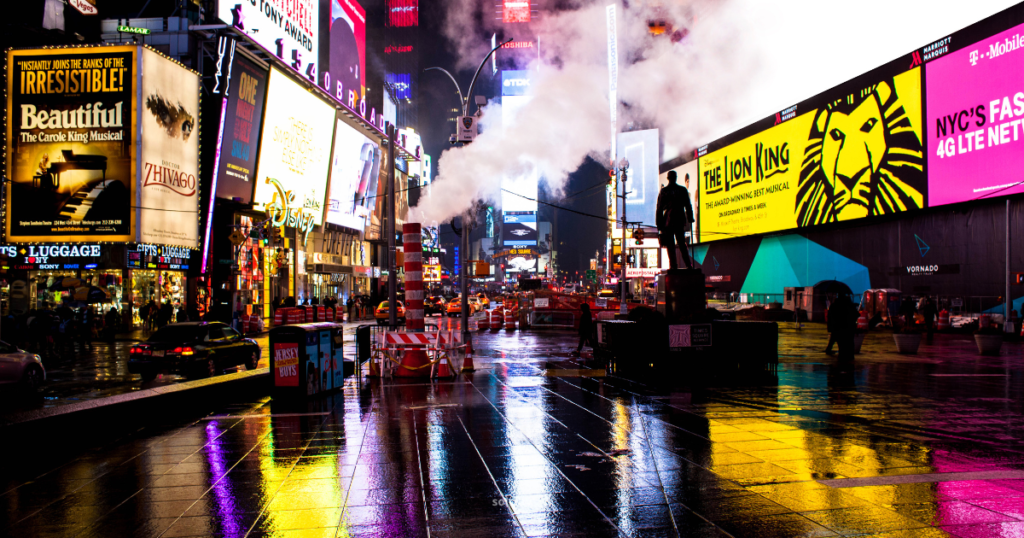 Nighttime view of Times Square, illuminated by vibrant neon lights and large digital billboards. The wet pavement reflects the colorful advertisements, creating a dynamic atmosphere. Steam rises from vents, adding to the urban ambiance, while a statue stands prominently in the foreground amidst the bustling scene.