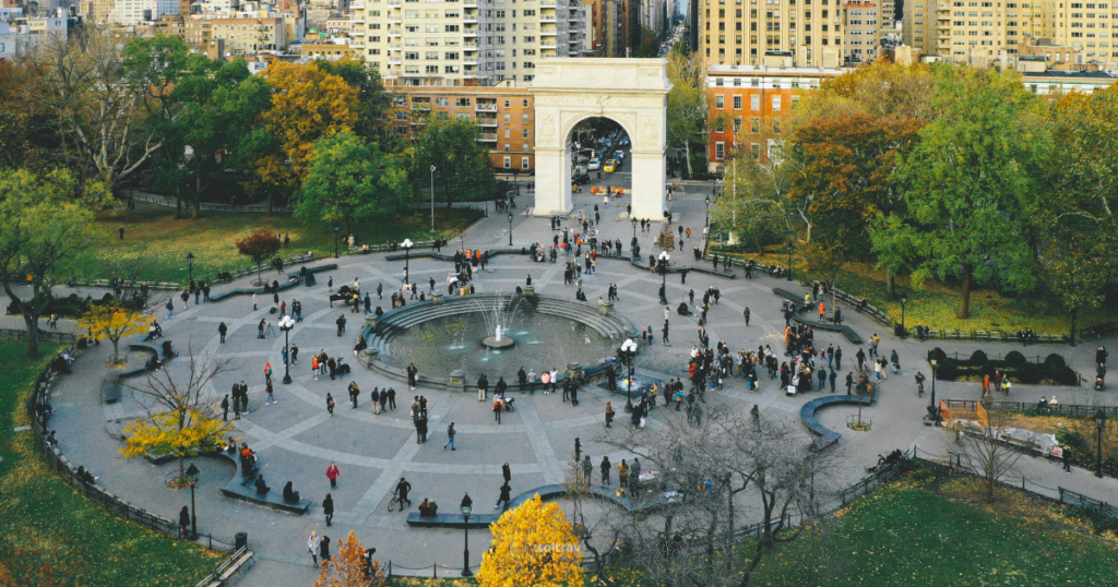 Aerial view of Washington Square Park in Lower Manhattan, New York City, showcasing the iconic Washington Arch in the background. The park is filled with visitors, some gathered around the central fountain, while others stroll along the pathways. Surrounding trees display autumn foliage in vibrant hues, creating a lively and inviting atmosphere.