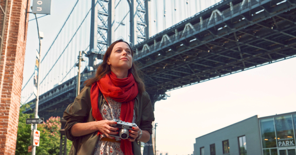 A young tourist standing on the Brooklyn Bridge, looking up with a thoughtful expression. She is holding a camera in her hands, dressed in a stylish outfit with a red scarf. The iconic bridge structure looms in the background, highlighting the architectural details of the cables and supports. The scene captures a sense of exploration and the beauty of the surroundings.