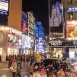 A vibrant street view of Myeongdong in Seoul, illuminated by colorful neon lights and large advertisements. Crowds of people stroll through the bustling shopping district, surrounded by shops and food stalls.