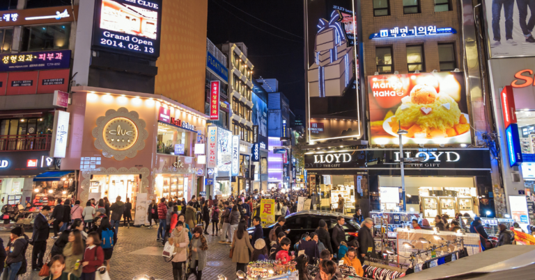 A vibrant street view of Myeongdong in Seoul, illuminated by colorful neon lights and large advertisements. Crowds of people stroll through the bustling shopping district, surrounded by shops and food stalls.