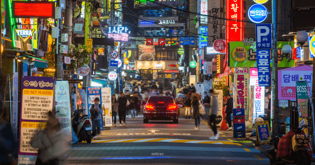 A lively street view of Sinchon in Seoul, illuminated by colorful neon signs. Crowds of people walk along the bustling street, surrounded by shops, restaurants, and vibrant advertisements.