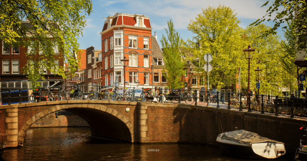 A picturesque scene in Amsterdam, Netherlands, featuring a charming canal, a small bridge, and traditional Dutch buildings. Bicycles are parked along the canal, and a boat is moored nearby.