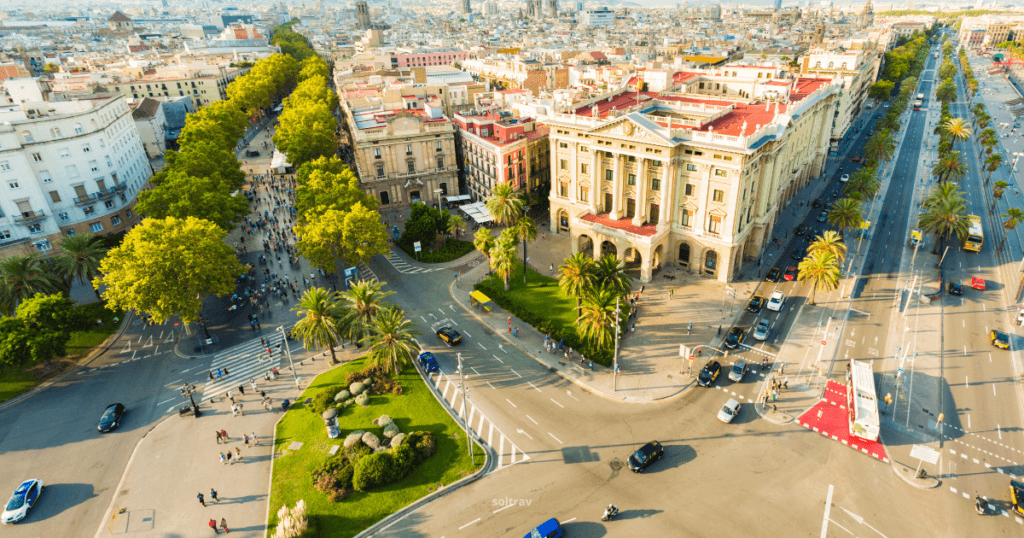 Aerial view of a bustling street in Barcelona, Spain, showcasing palm trees, green spaces, and historic buildings. The vibrant city life is visible with people walking and cars driving.