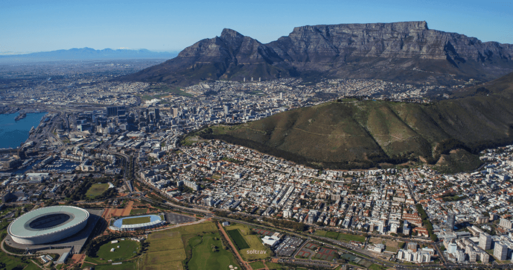 Aerial view of Cape Town, South Africa, showcasing Table Mountain and the city below. The image features the iconic stadium and the coastline, with lush greenery in the foreground.