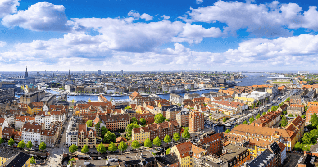 A panoramic view of Copenhagen, Denmark, showcasing colorful historic buildings along the waterfront, with canals and boats. The skyline features a mix of modern and traditional architecture under a partly cloudy sky.