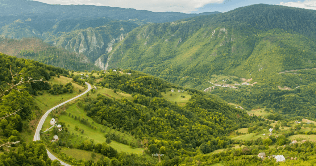 A panoramic view of Durmitor National Park in Montenegro, featuring lush green valleys, winding roads, and distant mountains under a cloudy sky.