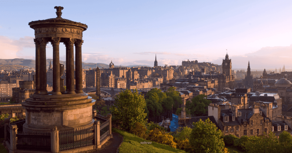A panoramic view of Edinburgh, Scotland, featuring historical buildings and the iconic Edinburgh Castle. A classical monument stands in the foreground, with the cityscape bathed in warm sunlight.