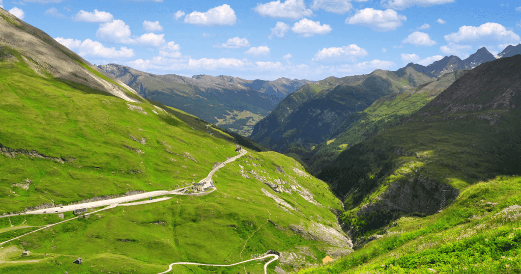 A panoramic view of Hohe Tauern National Park in Austria, featuring lush green valleys, winding roads, and majestic mountains under a blue sky with fluffy clouds.