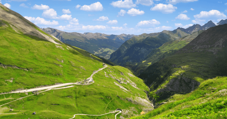 A panoramic view of Hohe Tauern National Park in Austria, featuring lush green valleys, winding roads, and majestic mountains under a blue sky with fluffy clouds.