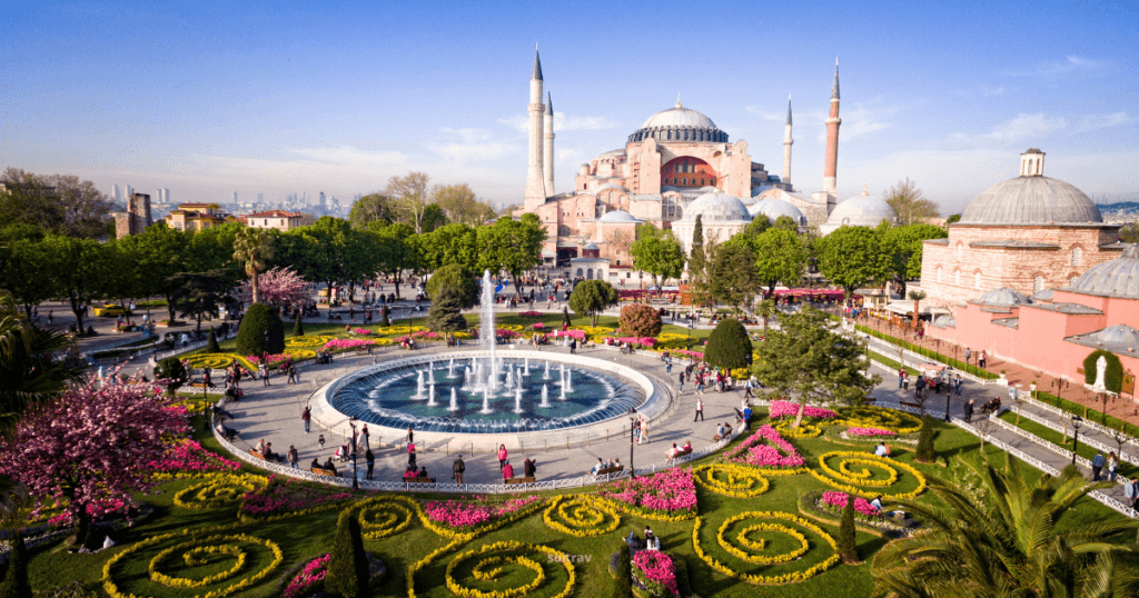 Aerial view of the Hagia Sophia in Istanbul, Turkey, surrounded by lush gardens and colorful flower beds. A fountain is in the foreground, and people are strolling through the area.