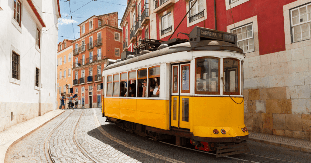 A historic yellow tram navigating a narrow street in Lisbon, Portugal. Colorful buildings line the street, and people can be seen enjoying the vibrant atmosphere.