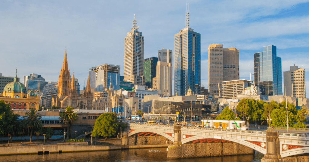 A stunning skyline of Melbourne, Australia, featuring a mix of historic and modern buildings. The Yarra River flows in the foreground, with a bridge connecting the two sides.