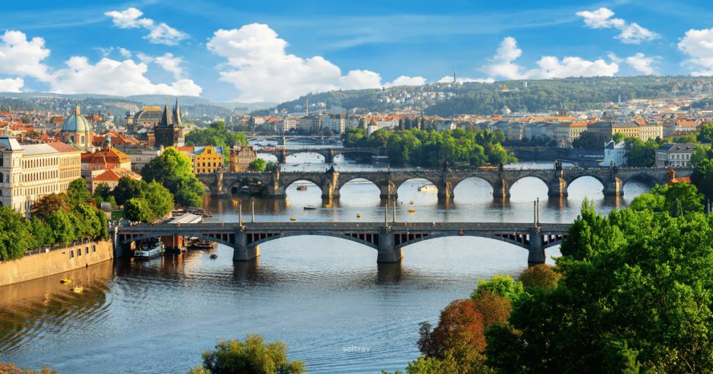 A panoramic view of Prague, Czech Republic, featuring the Vltava River and multiple historic bridges. The skyline includes colorful buildings and greenery under a bright blue sky with fluffy clouds.