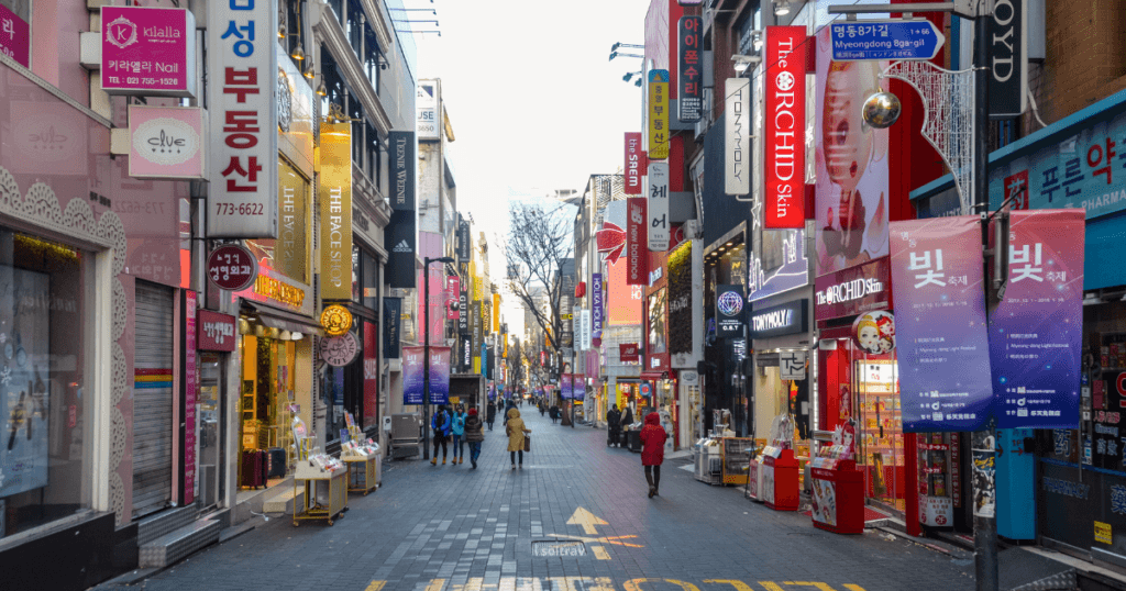 A bustling shopping street in Seoul, South Korea, lined with shops and colorful advertisements. People walk along the street, which is adorned with vibrant signage in Korean.