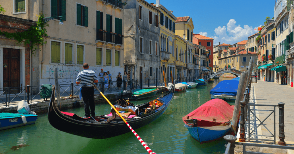 A picturesque canal in Venice, Italy, featuring a gondola with a gondolier and colorful boats along the water. Charming buildings line the canal under a bright blue sky.
