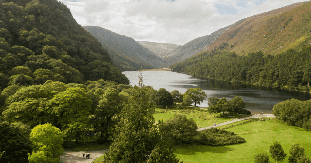 A scenic view of Wicklow Mountains National Park in Ireland, featuring a serene lake surrounded by lush green forests and rolling hills under a partly cloudy sky.