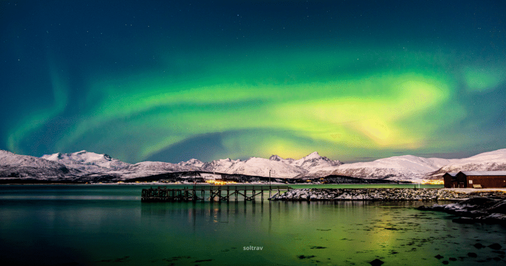 Stunning view of the Northern Lights over Tromsø, Norway, with snow-covered mountains and a calm fjord reflecting the vibrant colors.