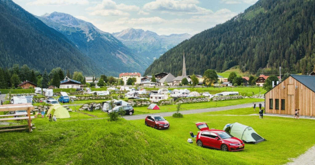 A scenic alpine campsite in Hohe Tauern, Austria, with colorful tents, camper vans, and a village surrounded by lush green mountains and distant glaciers.