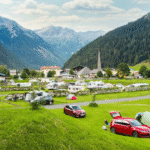 A scenic alpine campsite in Hohe Tauern, Austria, with colorful tents, camper vans, and a village surrounded by lush green mountains and distant glaciers.