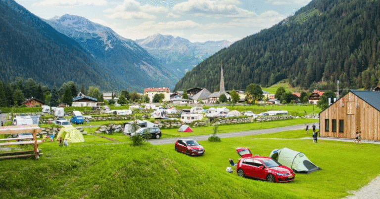 A scenic alpine campsite in Hohe Tauern, Austria, with colorful tents, camper vans, and a village surrounded by lush green mountains and distant glaciers.