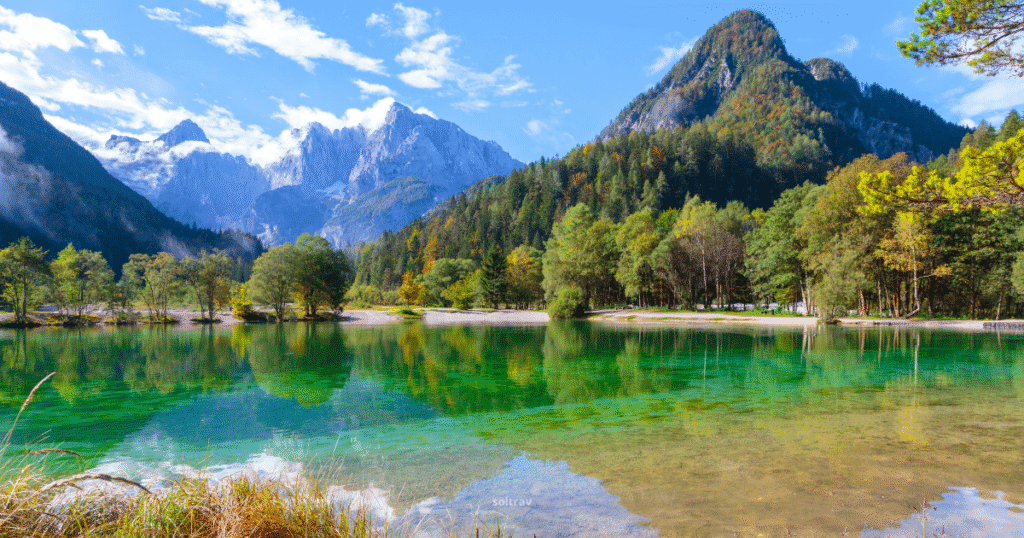 Crystal-clear emerald lake surrounded by trees and towering peaks of the Julian Alps in Slovenia under a bright blue sky.