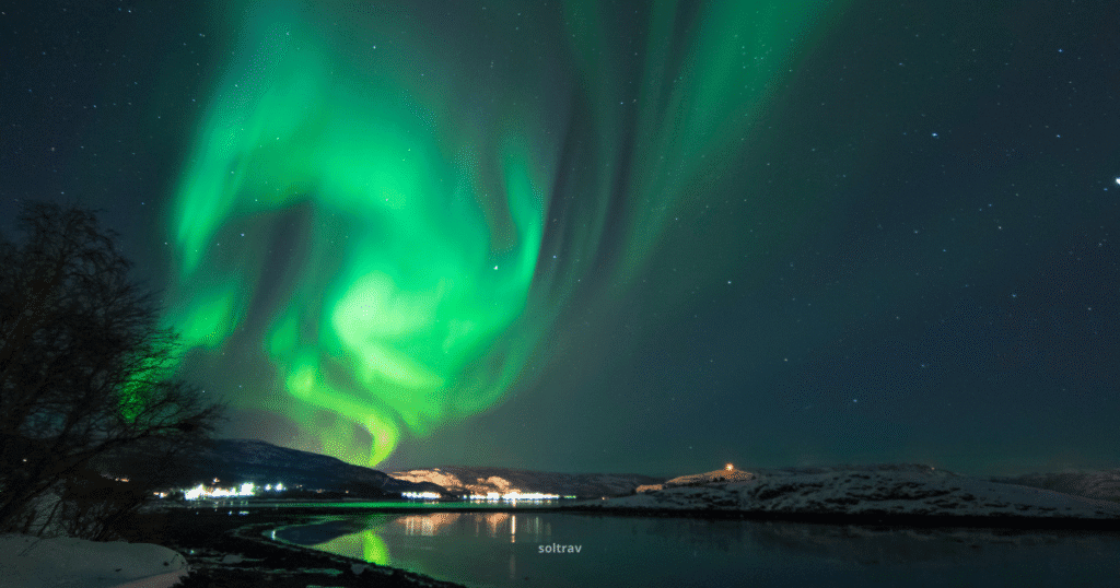Vibrant Northern Lights illuminate the sky over Alta, Norway, with a tranquil water reflection and snow-covered hills in the background.