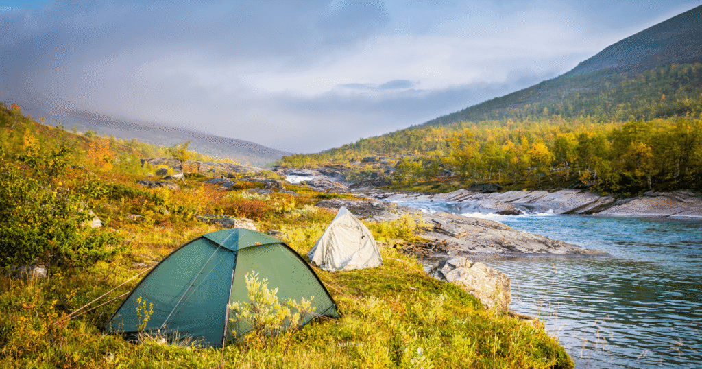 Two tents set up in the Arctic Circle wilderness of Lapland, Sweden, next to a clear river with autumn foliage on the surrounding hills.