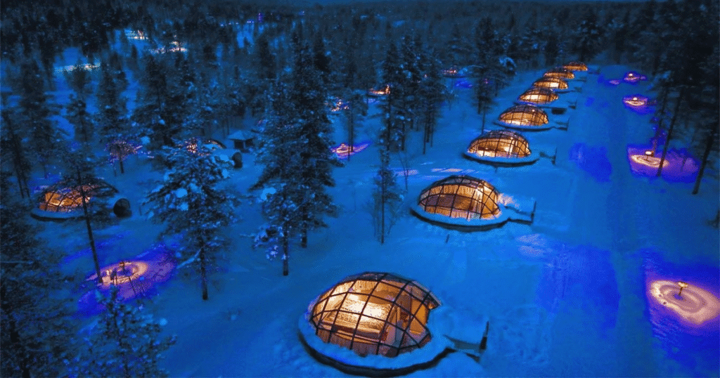Aerial view of glass igloos illuminated in Finnish Lapland, surrounded by snow-covered trees in a winter landscape.