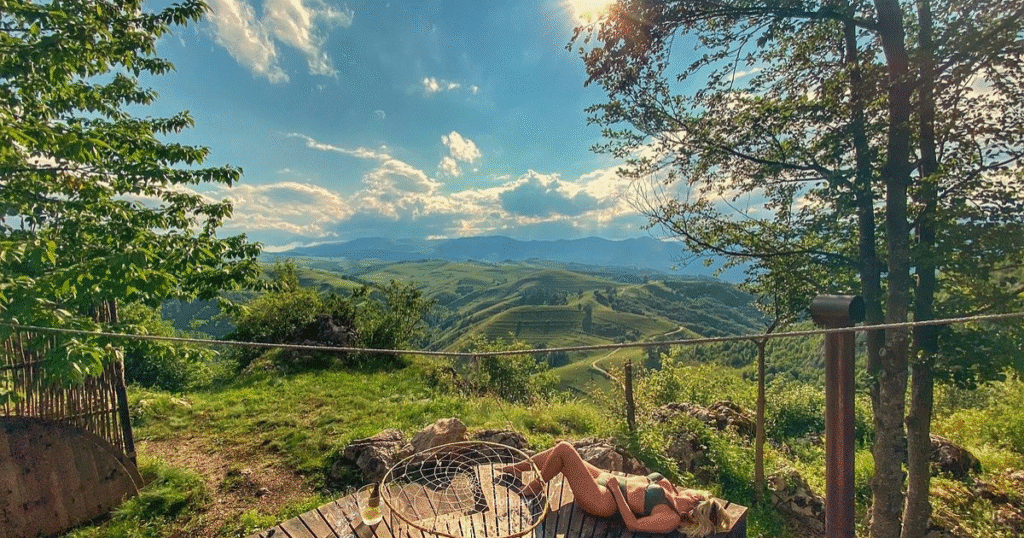 A woman relaxing on a wooden deck overlooking a scenic, rolling green valley in Transylvania, Romania, under a bright blue sky with scattered clouds.