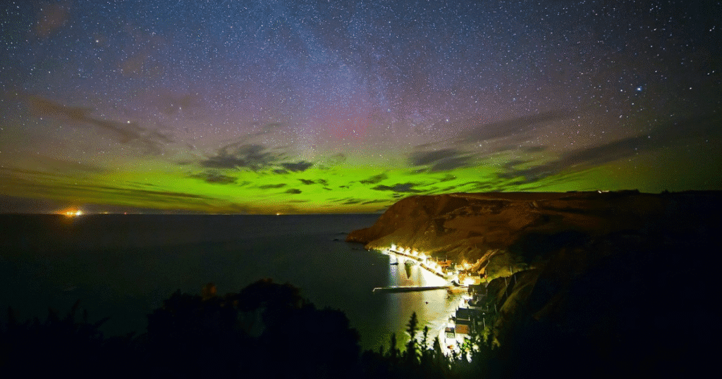 Enchanting Northern Lights over the Isle of Skye, Scotland, with a coastal village illuminated by lights under a starry sky.