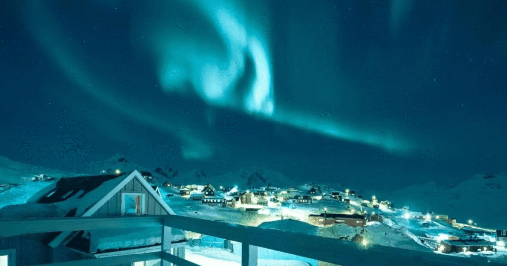 Stunning Northern Lights illuminate the night sky over Kangerlussuaq, Greenland, with a view of illuminated houses and snow-covered mountains.