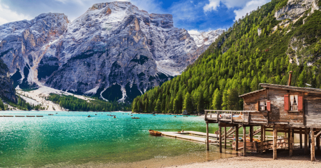 A rustic wooden cabin on stilts by the turquoise waters of Lago di Braies, surrounded by pine forests and towering peaks of the Dolomites, Italy.