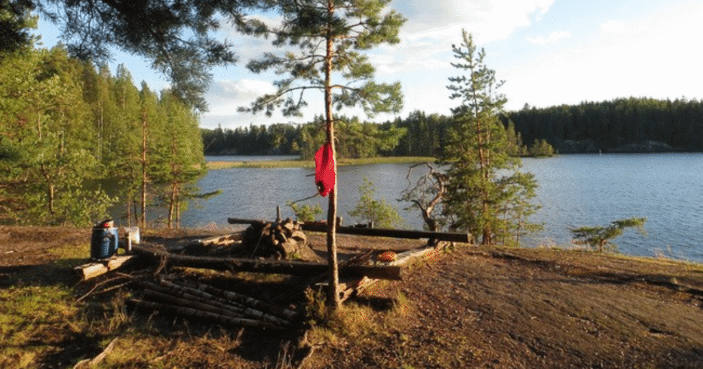 A quiet lakeside campsite in Saimaa, Finland, with a firepit, scattered logs, and a red shirt hanging from a pine tree beside a calm forest lake.