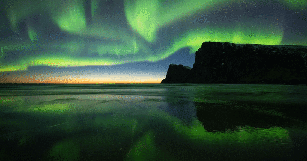 Breathtaking Northern Lights over the Lofoten Islands in Norway, reflecting on the calm waters with rugged cliffs in the background.