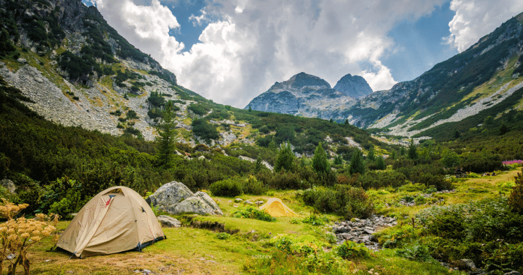 Scenic mountain landscape in the Rila Mountains, Bulgaria, featuring two tents nestled in a green valley surrounded by towering peaks and a cloudy sky.