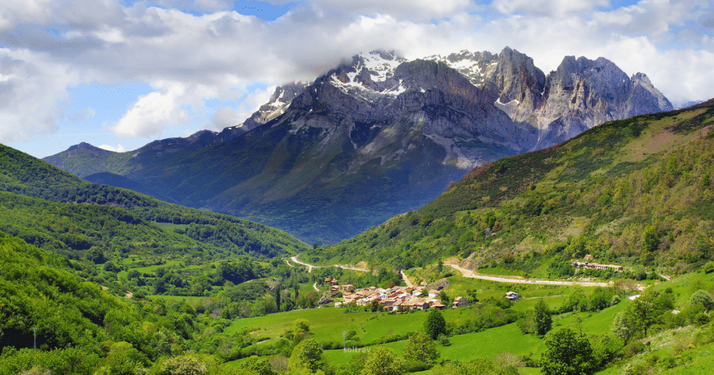 A picturesque mountain village nestled in a lush green valley with towering, snow-dusted peaks in the background in the Picos de Europa, Spain.