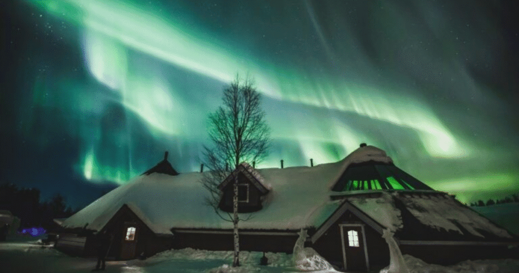 Vibrant Northern Lights above a snow-covered lodge in Rovaniemi, Finland, with trees silhouetted against the night sky.