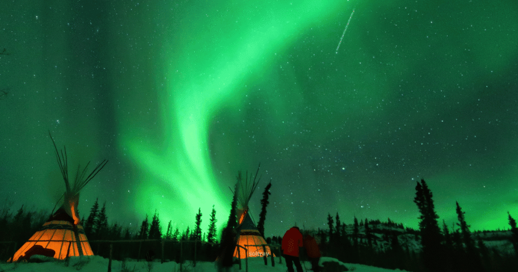 Vibrant Northern Lights illuminate the sky over Yukon Territory, Canada, with illuminated teepees and silhouetted figures in the foreground.